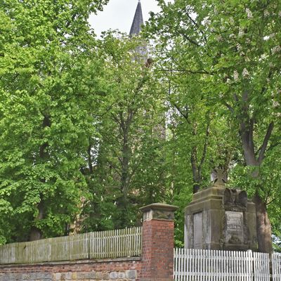 Ein Blick aus nord-westlicher Richtung zum Kirchengelände. Während der 42 Meter hohe Kirchturm durch die stattlichen Bäume schimmert, ist im Vordergrund ein Denkmal zur Erinnerung der in Kriegen gefallenen Ausleber zu sehen.