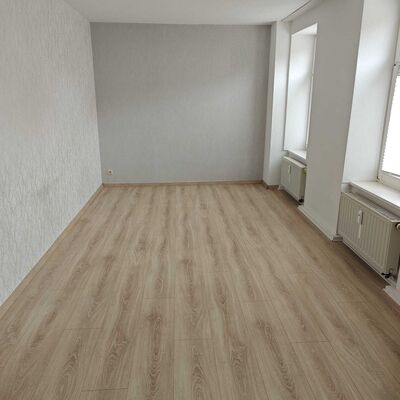 Das Bild zeigt das helle Wohnzimmer der 3-Raum-Wohnung in der Stra&szlig;e An der Tr&auml;nke 4 in 39397 Kroppenstedt. Der gro&szlig;z&uuml;gig geschnittene Wohnraum mit modernem Bodenbelag bietet viel Platz f&uuml;r individuelles Wohnen und schafft ein angenehmes Wohngef&uuml;hl. Die Wohnung liegt in ruhiger Lage in Kroppenstedt in der Verbandsgemeinde Westliche B&ouml;rde und eignet sich ideal f&uuml;r Paare, kleine Familien oder Menschen, die komfortables Wohnen im Landkreis B&ouml;rde suchen.